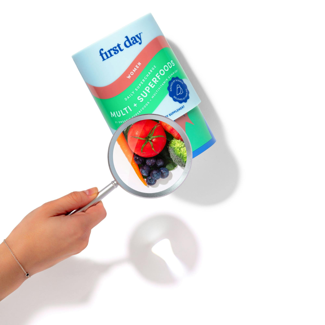 A hand holds a magnifying glass over a bottle labeled first day Multi + Superfoods, highlighting an image of tomatoes, blueberries, carrots, and broccoli on the bottle.