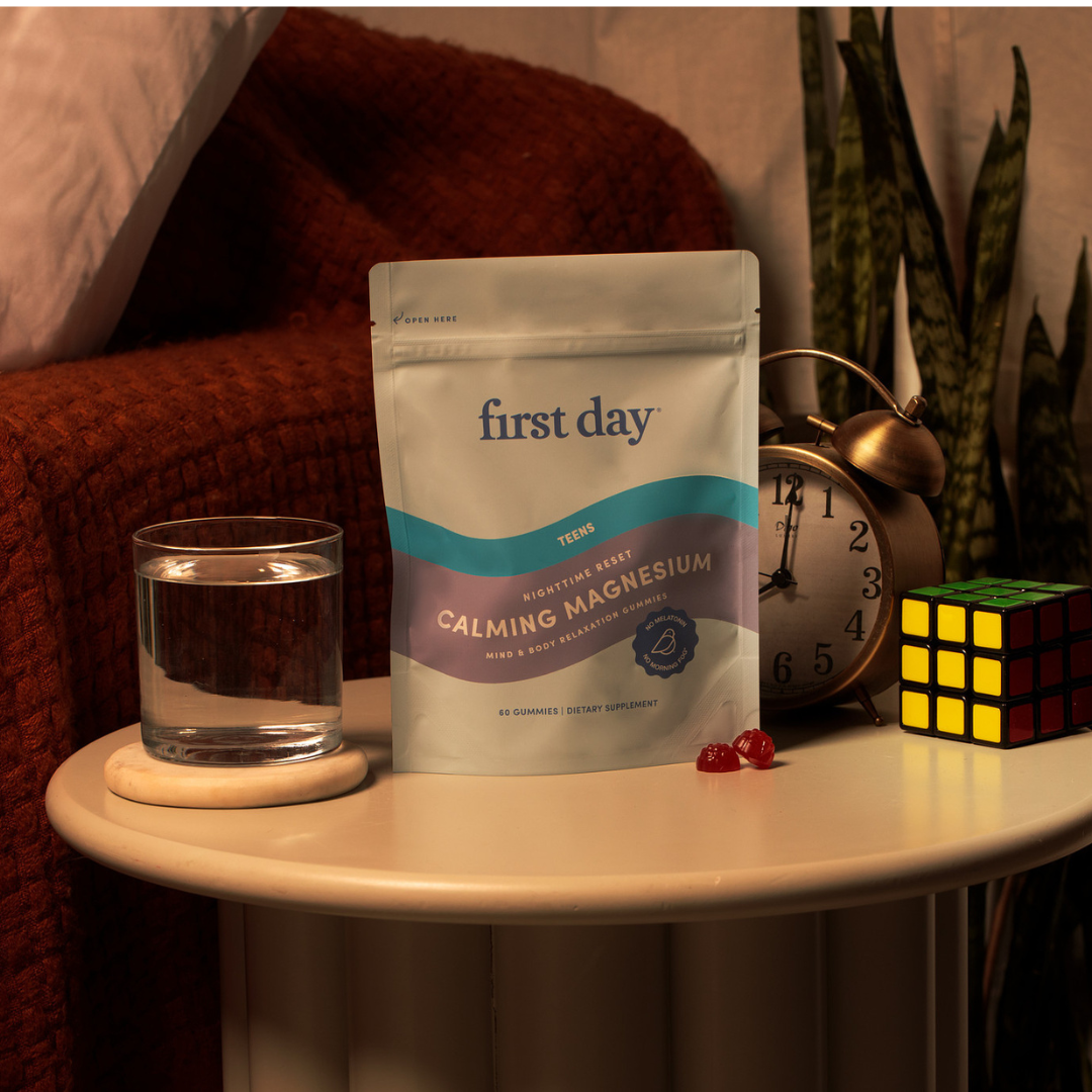 A pouch of First Day Calming Magnesium Teen gummies sits on a round bedside table with a glass of water, two red gummies, a classic alarm clock, a Rubiks cube, and a textured rust-colored blanket in the background.