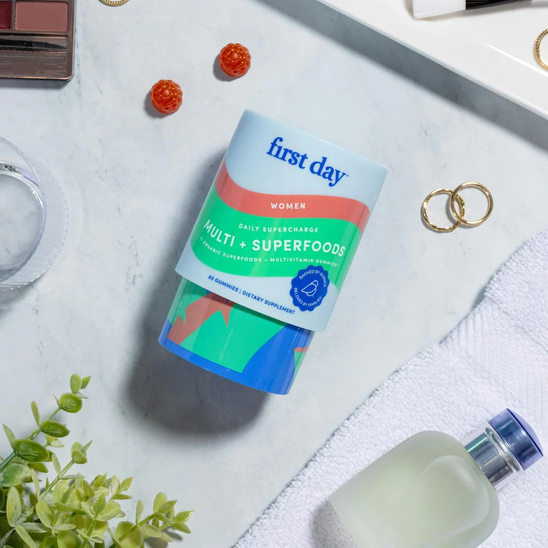 A container of First Day Women’s Multi + Superfoods vitamins sits on a marble surface surrounded by gold rings, a white towel, a bottle of perfume, a glass, and two orange gummies.