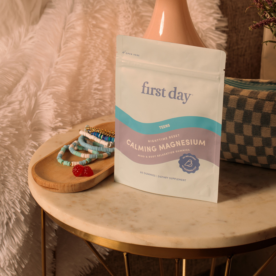 A pouch labeled “first day Calming Magnesium” sits on a round marble table beside colorful beaded bracelets, a red gummy, a lamp, and a chevron-patterned pillow, with a fuzzy white blanket in the background.