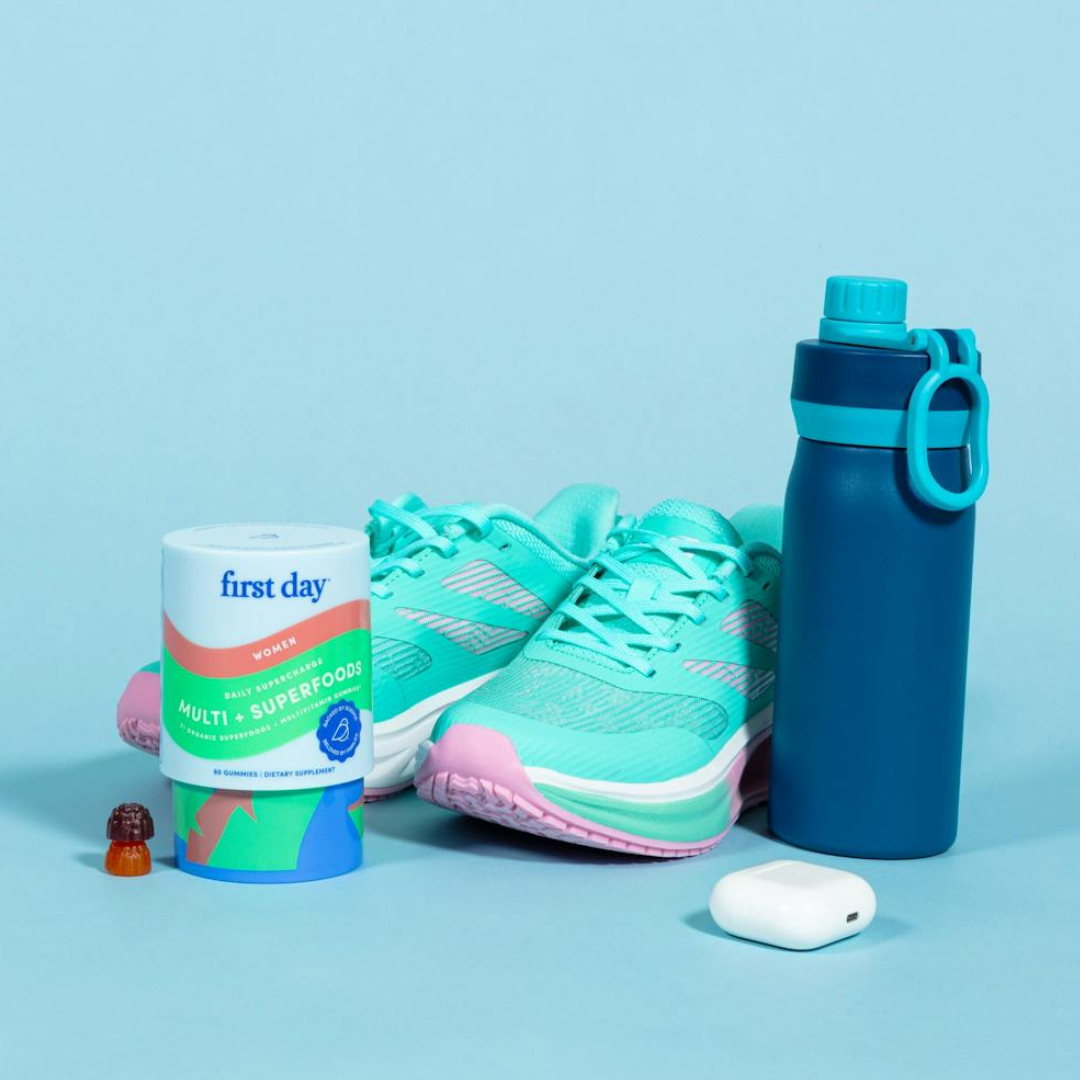 A pair of teal running shoes, a blue water bottle, a container of “first day” multivitamins, a single vitamin gummy, and white wireless earbuds are arranged on a light blue background.