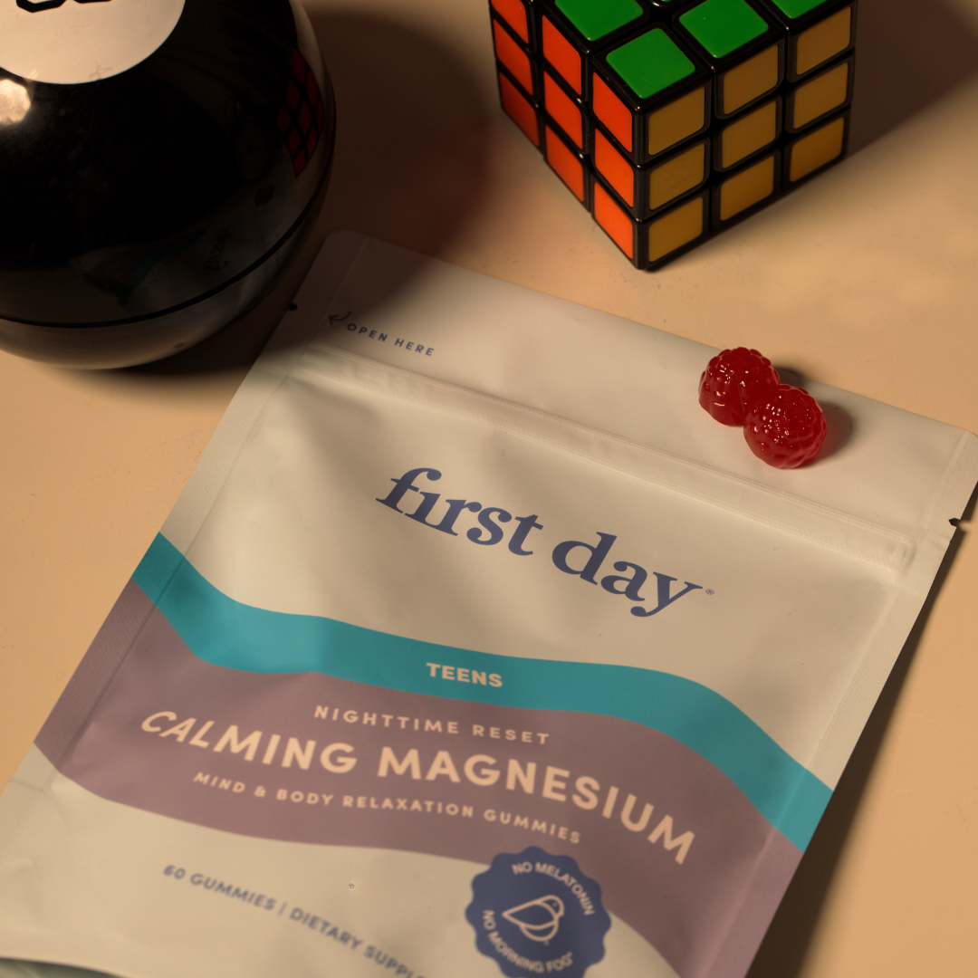 A pouch labeled first day Calming Magnesium Gummies lies on a table beside a Rubiks Cube, two red gummy candies, and a black object. The packaging is mostly white with blue and purple accents.