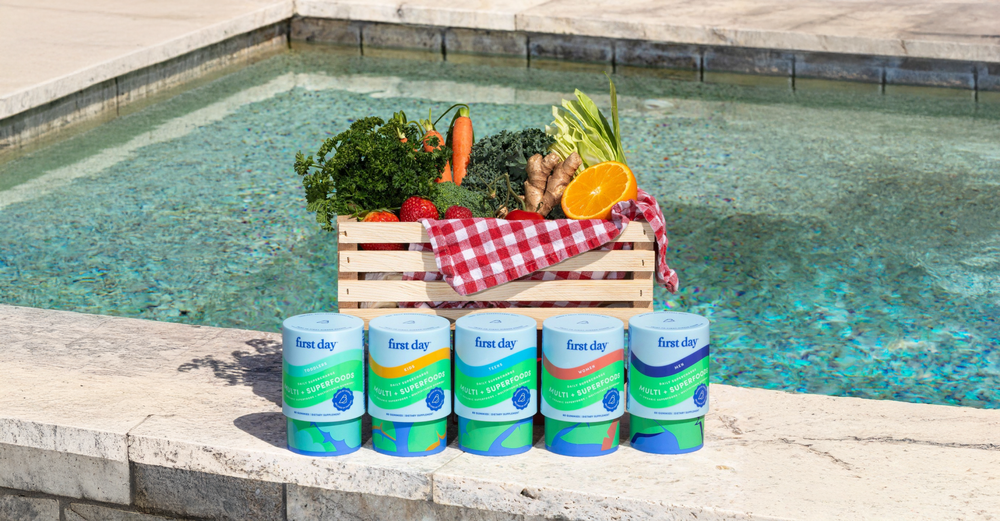 A wooden crate filled with fresh fruits and vegetables sits by a pool, with a red checkered cloth draped inside. In front, five First Day multivitamin bottles are lined up on the pool’s edge.