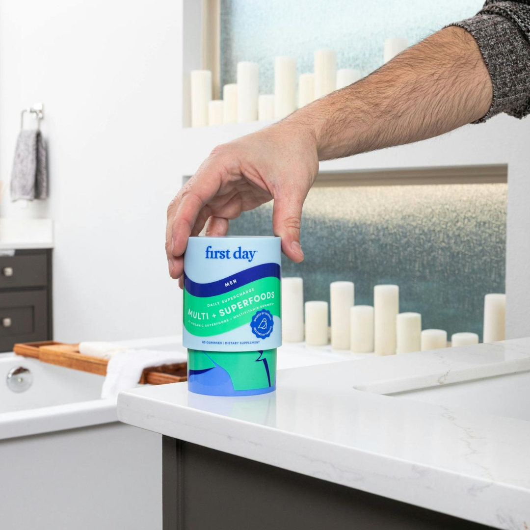 A person’s hand is reaching for a container labeled “first day Multi + Superfoods” on a white bathroom counter, with a bathtub, white rolled towels, and decorative candles in the background.
