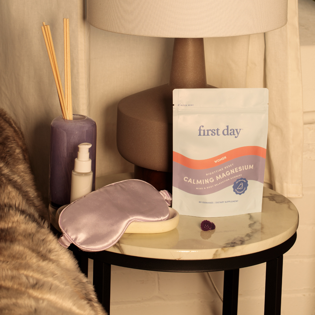 A bedside table with a lamp, reed diffuser, lotion, a sleep mask, and a pouch labeled first day Women’s Calming Magnesium supplement, along with a small purple stone, next to a furry blanket.