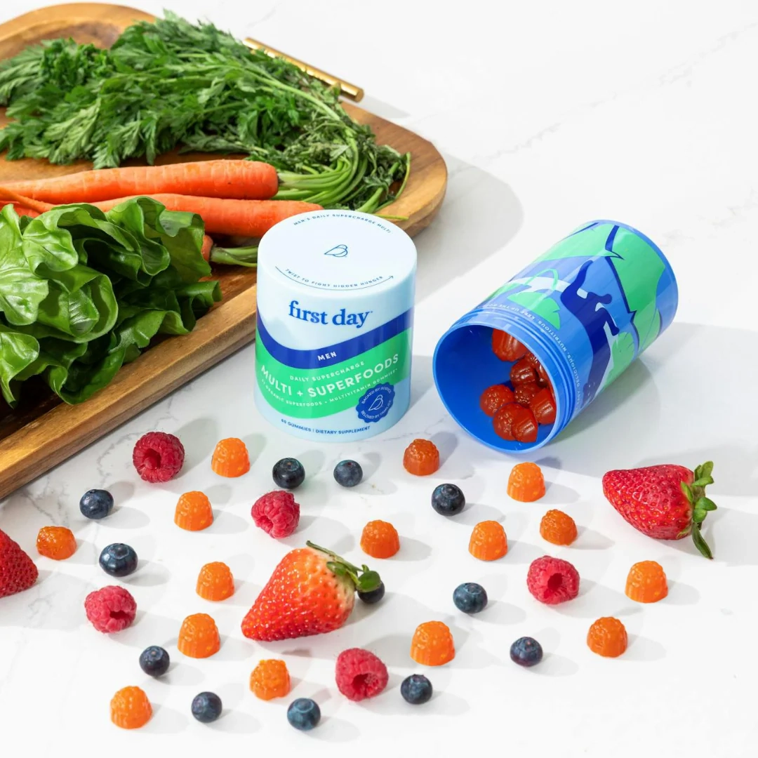 A wooden tray with leafy greens and carrots sits beside two supplement containers on a white surface, surrounded by scattered berries, strawberries, and orange gummy vitamins.