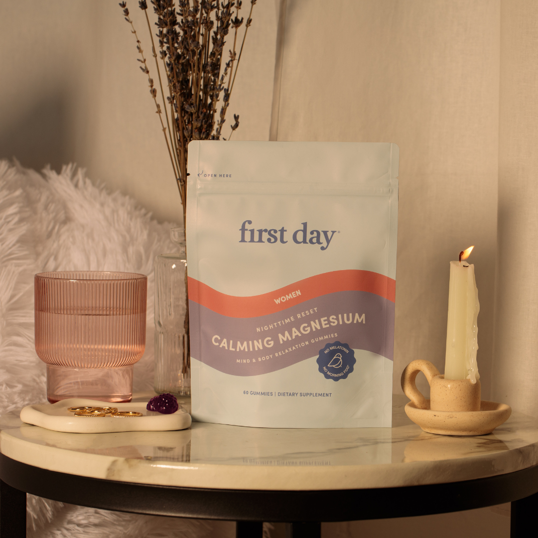 A pouch of First Day Calming Magnesium gummies sits on a round marble table next to a pink glass, a lit candle in a holder, a dish with jewelry, and a vase of dried flowers, creating a cozy, relaxing scene.