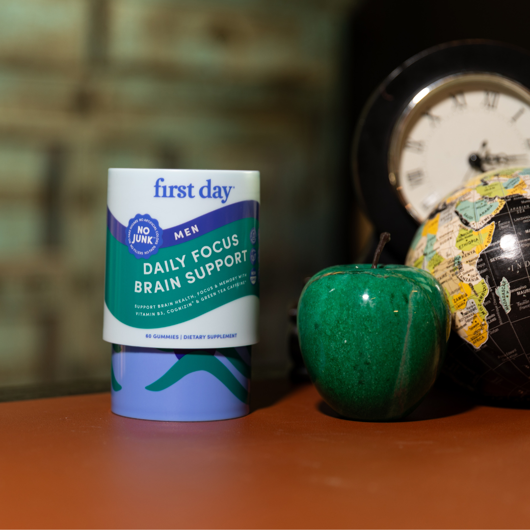 A container of First Day Daily Focus Brain Support gummies for men is displayed on a table next to a green apple, a small globe, and a clock in the background.