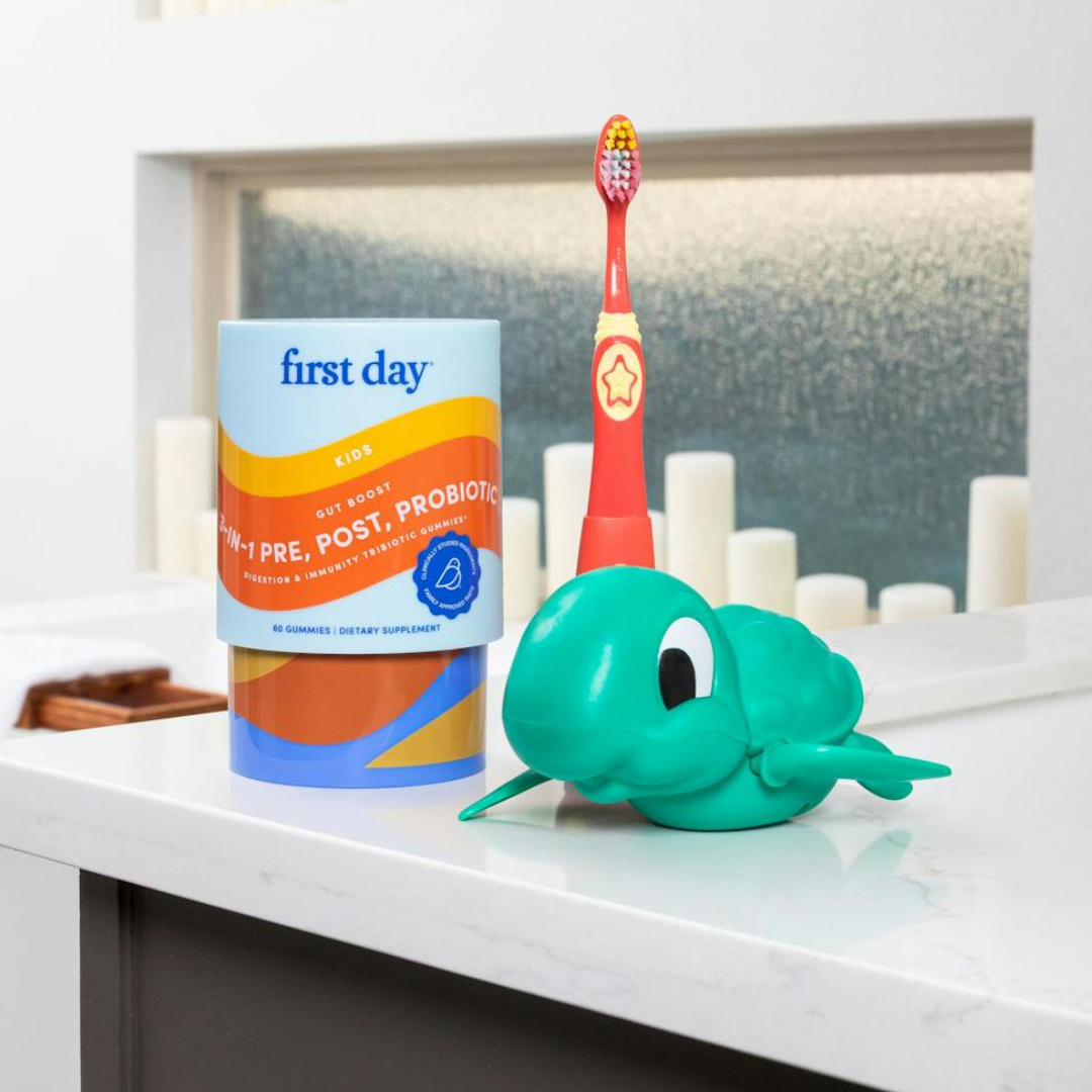A container of kids probiotics, a red and yellow kids’ toothbrush, and a green turtle-shaped object are placed on a white bathroom counter with candles in the background.