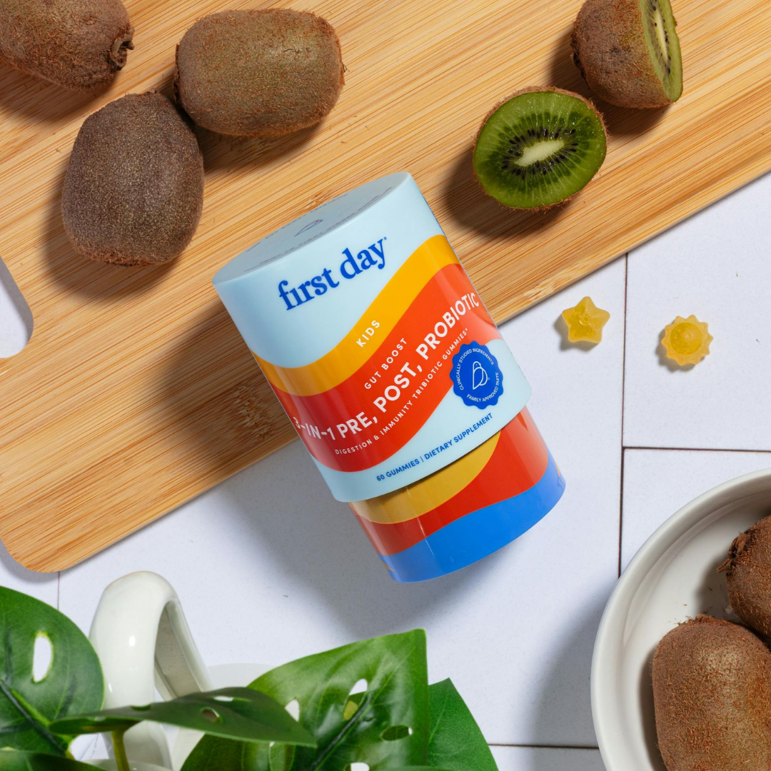 A container of First Day Kids Daily Probiotics lies on a wooden cutting board surrounded by whole and halved kiwis, two yellow star-shaped gummies, a plant, and a white mug on a white tiled surface.