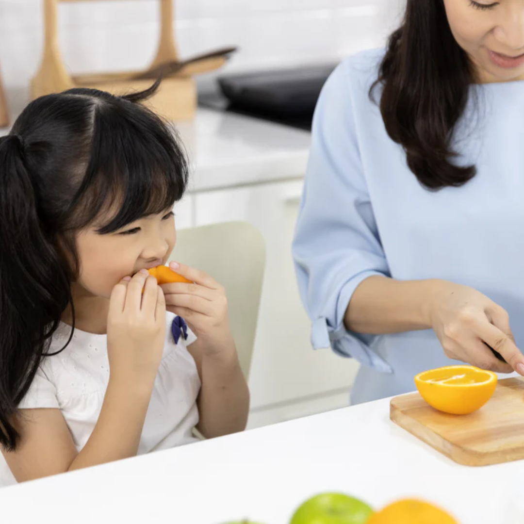 A young girl smiles as she eats an orange slice while an adult beside her slices another orange on a cutting board in a bright kitchen.