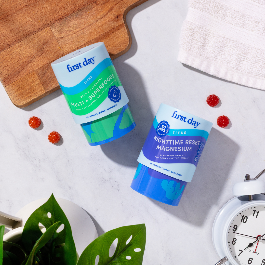 Two bottles from the Teens' Dream Team Starter Pack rest on a white surface near a plant, wooden board, towel, clock, and gummy vitamins—ideal for supporting teen multivitamin needs.