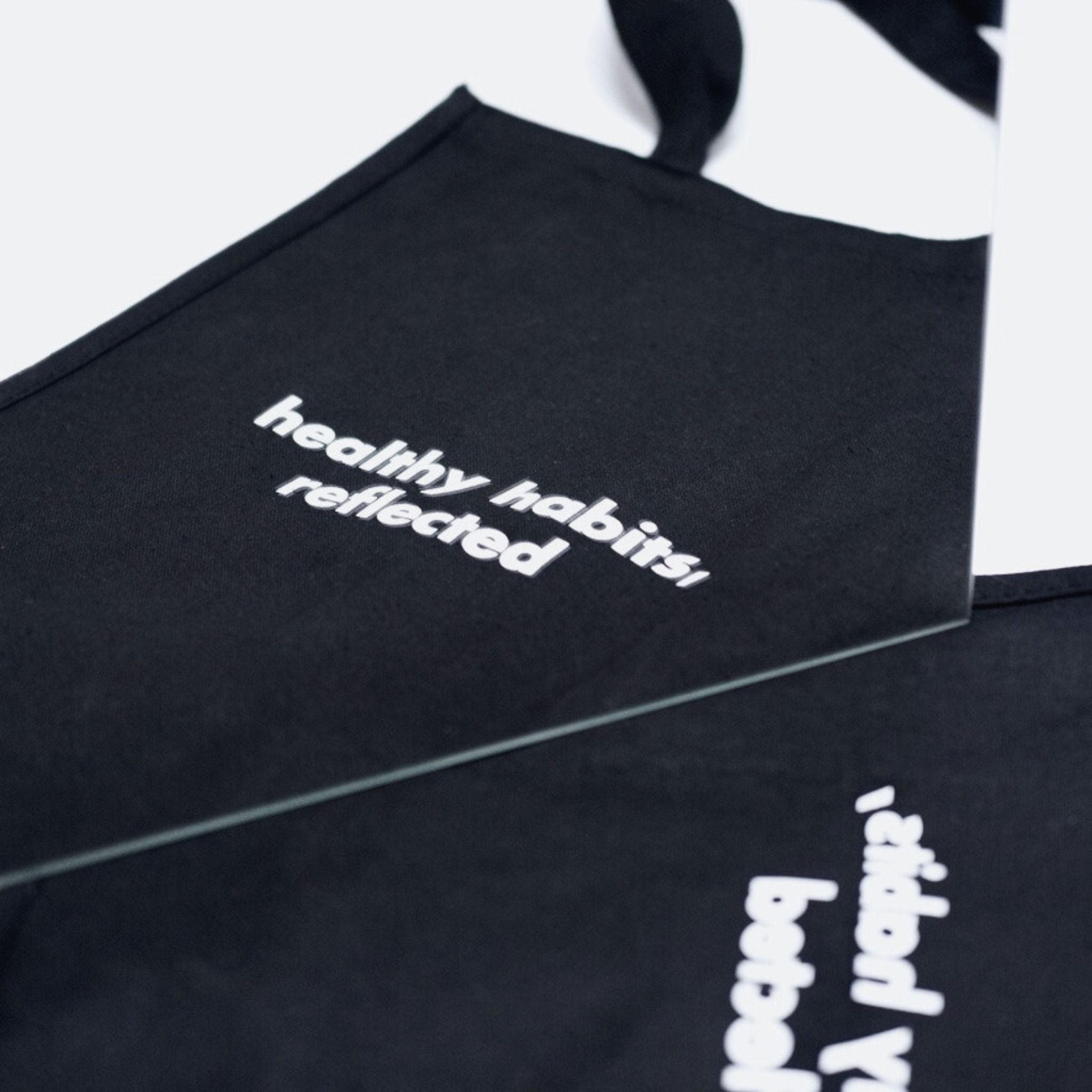 A black apron with "healthy habits, reflected" printed in white sits beside a mirror, echoing the mindful inspiration of the Reflection Tote.
