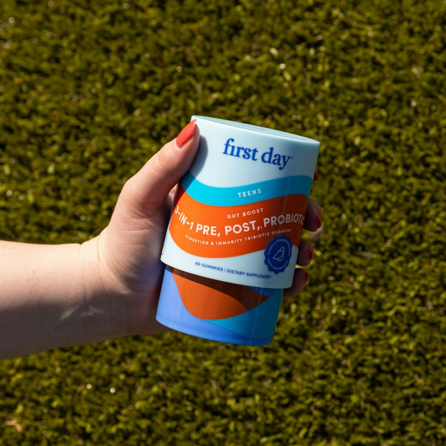 A hand with red nail polish holds a container labeled first day with 4-IN-1 PRE, POST, PROBIOTIC written on it, against a blurred green grass background.