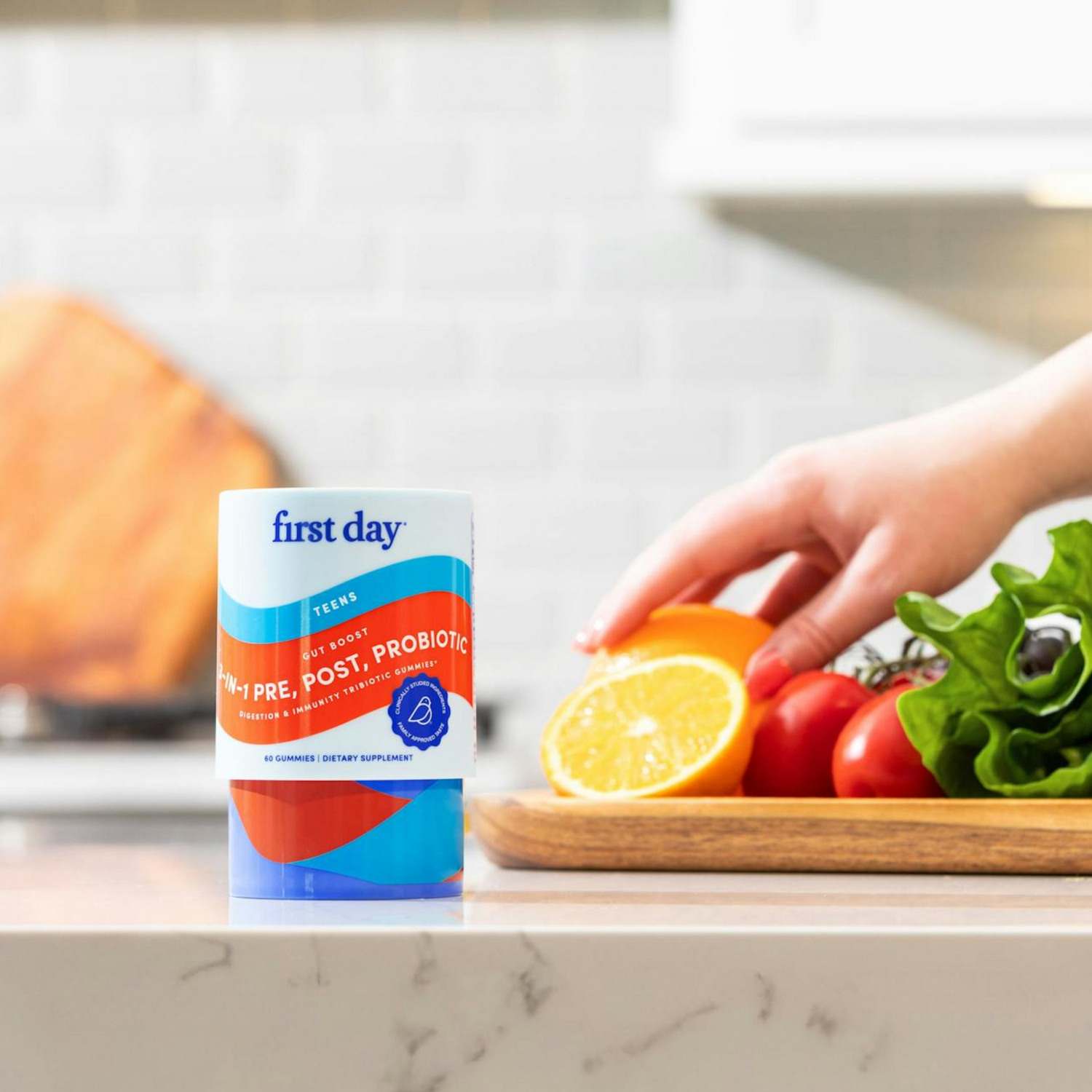 A hand holds a sliced lemon next to fresh tomatoes and lettuce on a wooden board, with a First Day teen pre, post, probiotic supplement container on a kitchen counter in the foreground.