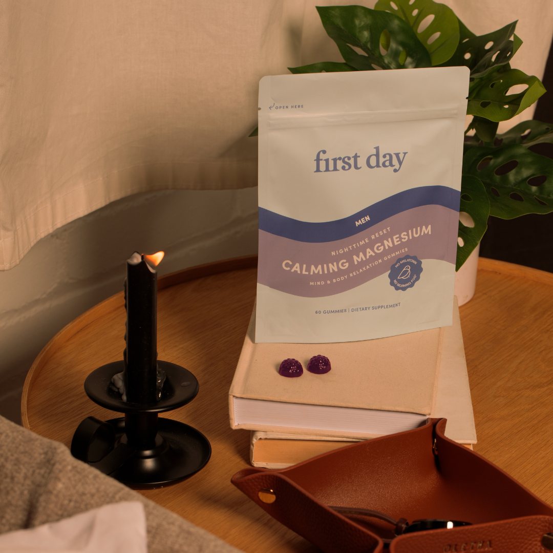 A packet labeled first day Calming Magnesium sits on a closed book next to two purple gummies, a lit black candle, and a leather tray on a wooden table with a green plant in the background.