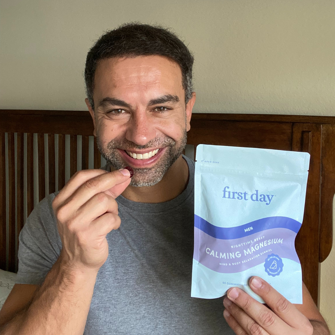 A smiling man in a gray shirt holds up a gummy supplement and a pouch labeled first day Men Calming Magnesium while sitting on a wooden bed.
