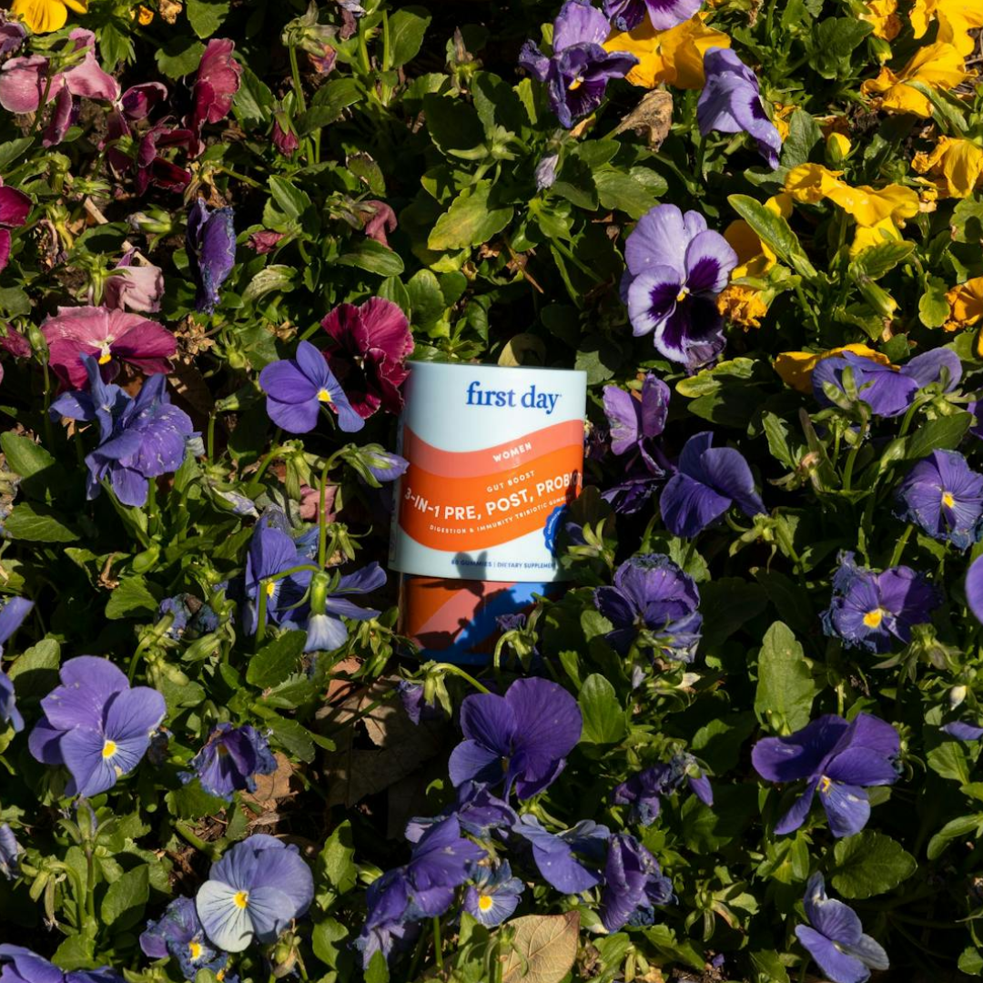 A container labeled first day Womens 3-in-1 Pre, Post, Probiotic is nestled among vibrant purple, yellow, and maroon pansies in a garden.