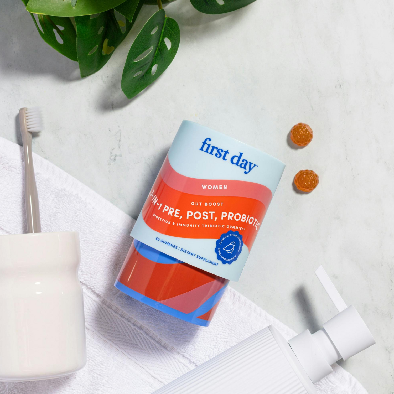 A container of First Day women’s pre, post, probiotic supplements lies on a marble surface near two gummies, a toothbrush in a holder, a white towel, a soap dispenser, and green leaves.
