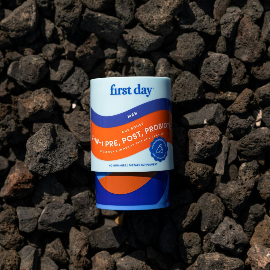 A container labeled first day with 4-in-1 Pre, Post, Probiotic for men rests on dark, rough volcanic rocks. The packaging is blue and orange with a modern design and contains 60 gummies.