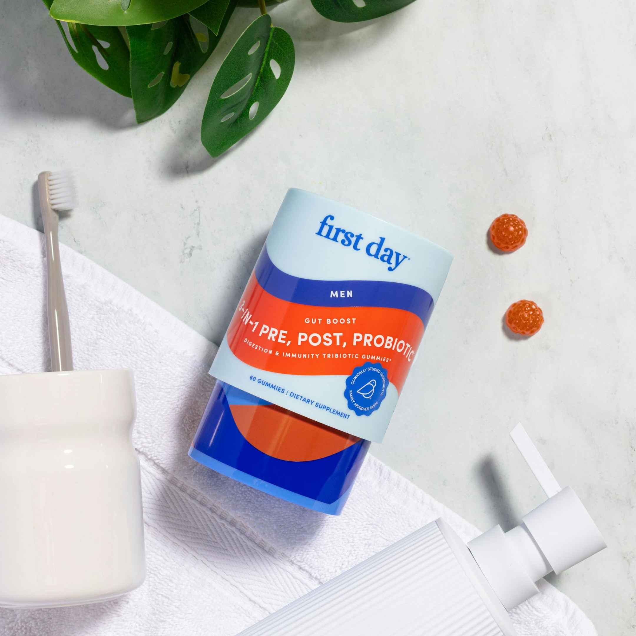 A container of First Day mens pre, post, and probiotic gummies lies on a towel next to a white cup holding a toothbrush, a soap dispenser, two gummies, and green leaves.
