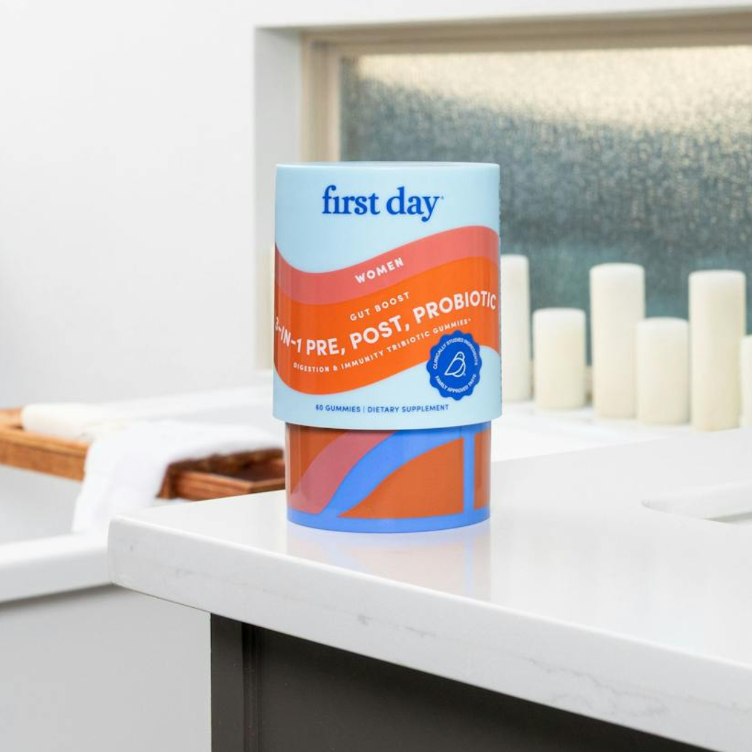 A container of First Day Women’s Gut Boost Pre, Post, Probiotic gummies sits on a bathroom counter near a bathtub and candles.