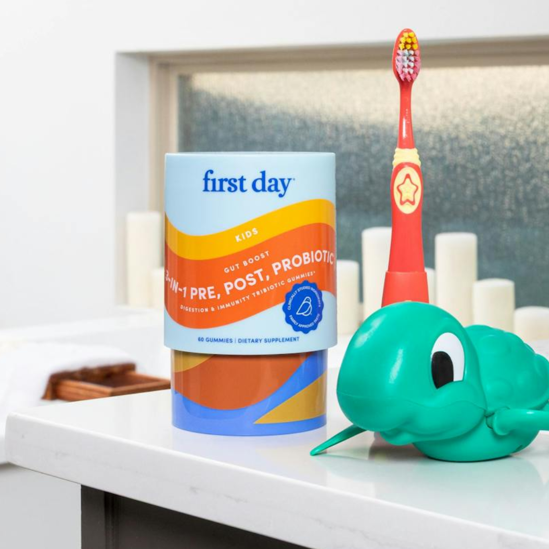 A container of First Day kids’ 3-in-1 pre, post, and probiotic gummies sits on a bathroom counter next to a childrens toothbrush with a star on it and a green turtle-shaped holder.