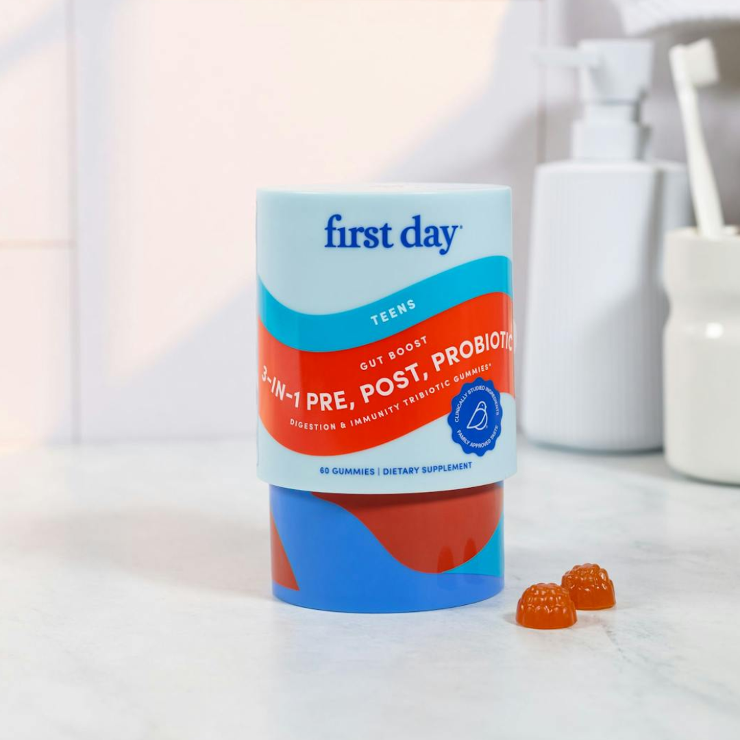 A container of First Day 2-in-1 Pre, Post, Probiotic gummies for teens is on a white countertop with two orange gummies beside it. Bathroom items are blurred in the background.