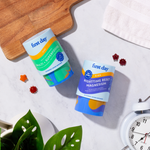 Two containers from the Kids' Dream Team Starter Pack sit on a marble surface near a clock, plant, towel, wooden board, and colorful organic superfood gummies.