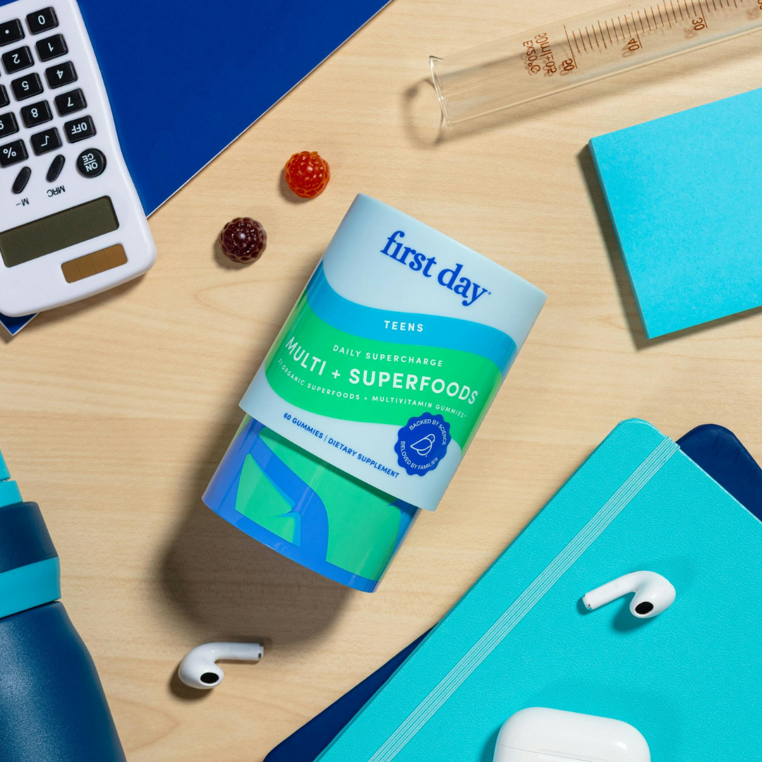 A container of First Day Teens Multi + Superfoods vitamins is surrounded by school supplies, including a calculator, notebook, sticky notes, pen, water bottle, AirPods, and gummy vitamins on a light blue surface.