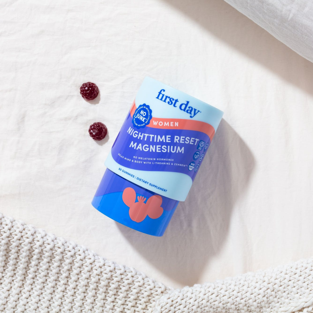 A bottle of First Day Women Nighttime Reset Magnesium gummies lies on a white surface next to two red gummies and a textured cream knit blanket.