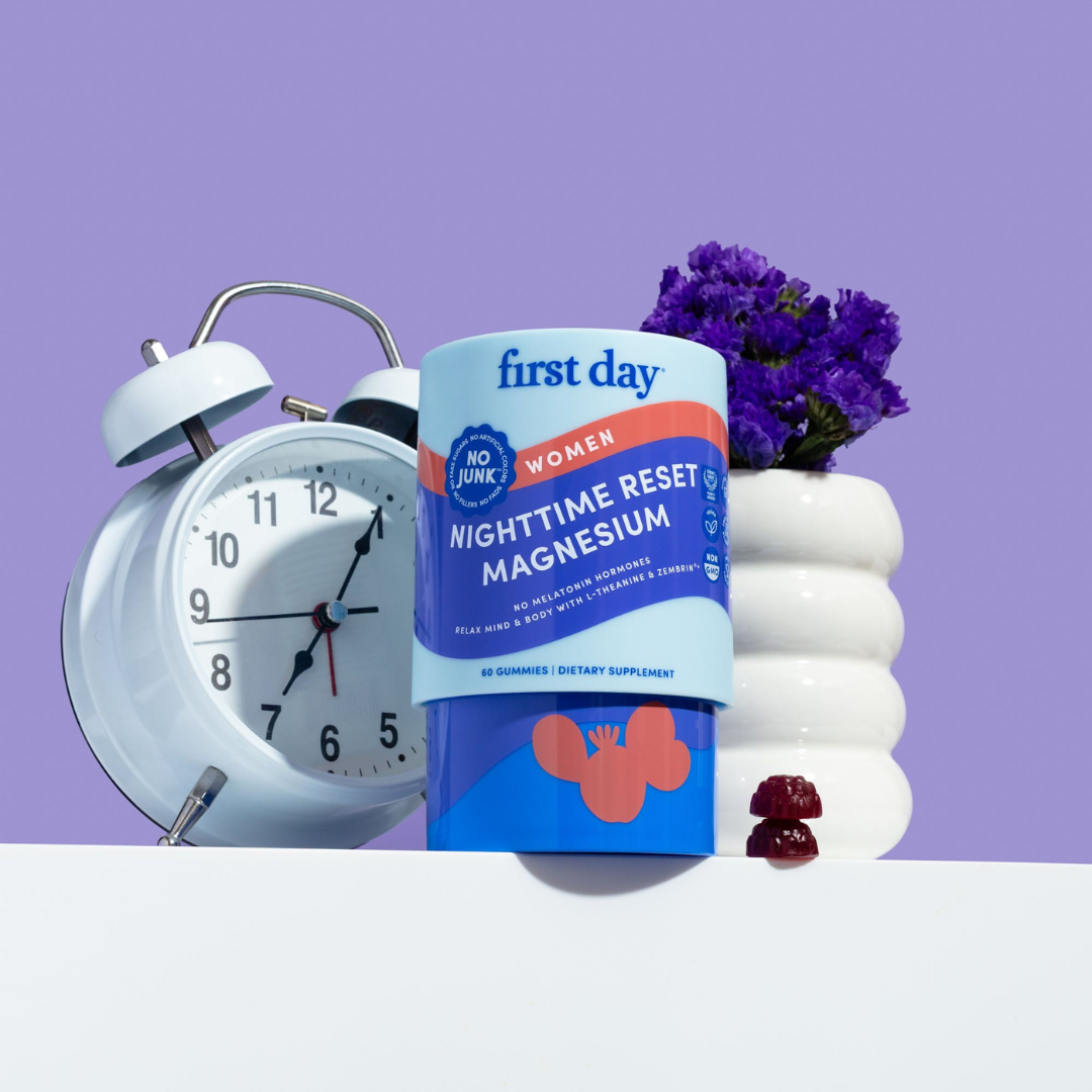 A blue bottle of First Day Nighttime Reset Magnesium gummies stands in front of a white alarm clock and next to a white vase with purple flowers, all set against a light purple background. Two gummies are placed beside the bottle.