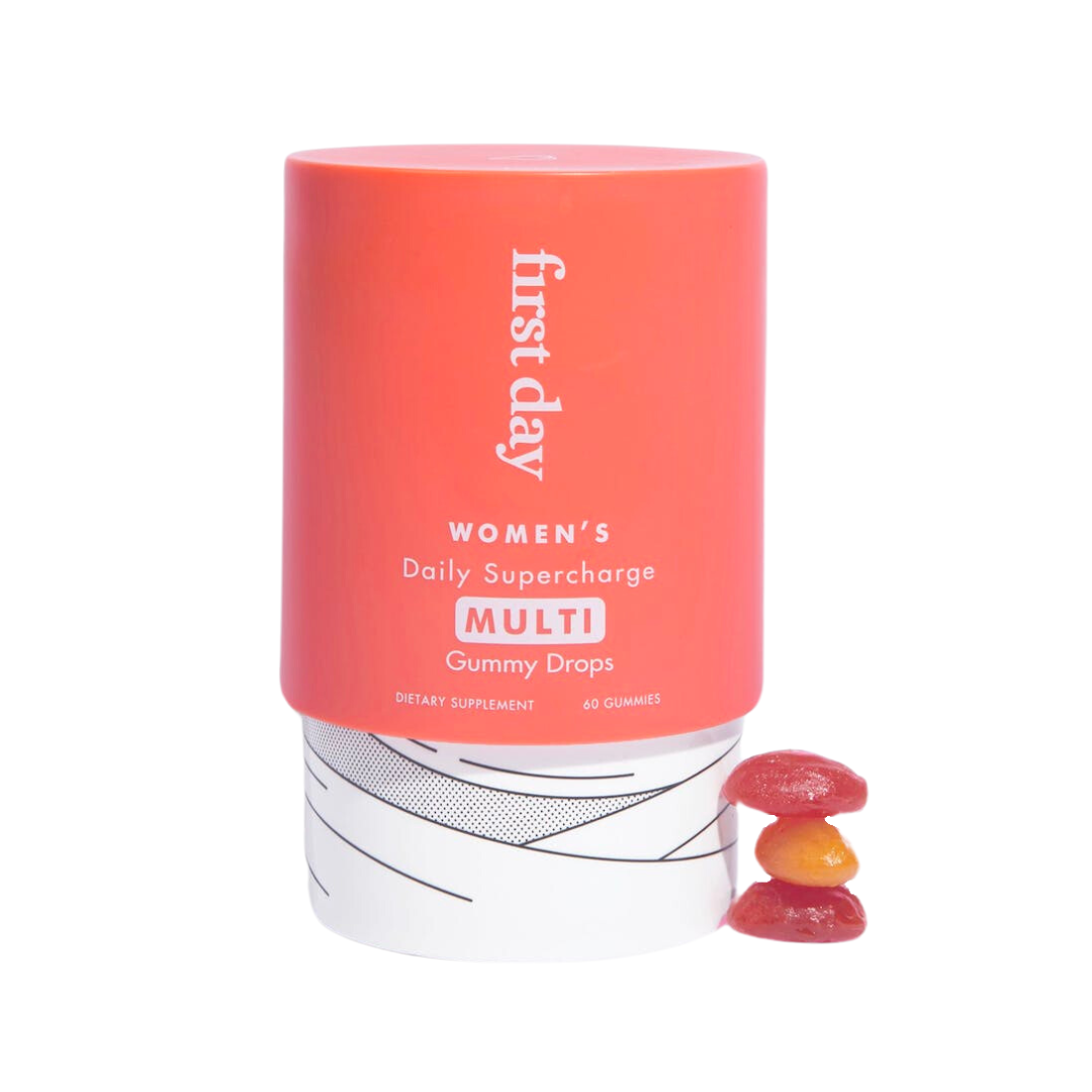 A container labeled first day Womens Daily Supercharge Multi Gummy Drops sits on a white surface, with three stacked, colorful gummy supplements placed in front of it.