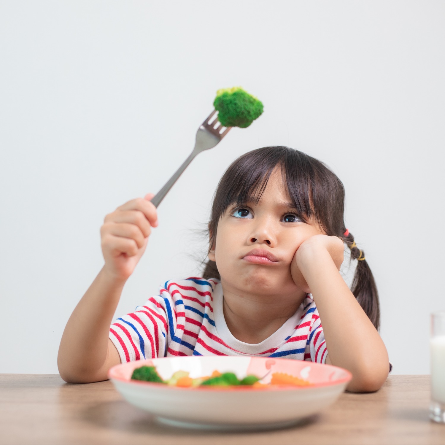 A young girl in a striped shirt sits at a table, resting her chin on her hand and looking bored while holding up a fork with a piece of broccoli. A plate of vegetables and a glass of milk are in front of her.