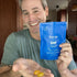 A smiling man holds three yellow gummy vitamins in one hand and a blue pouch labeled “first day men’s daily nutrition multi gummy” in the other, standing in a kitchen with brown cabinets in the background.