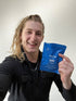 A smiling person with long blonde hair, wearing a black jacket, holds up a blue packet labeled first day against a plain white background.