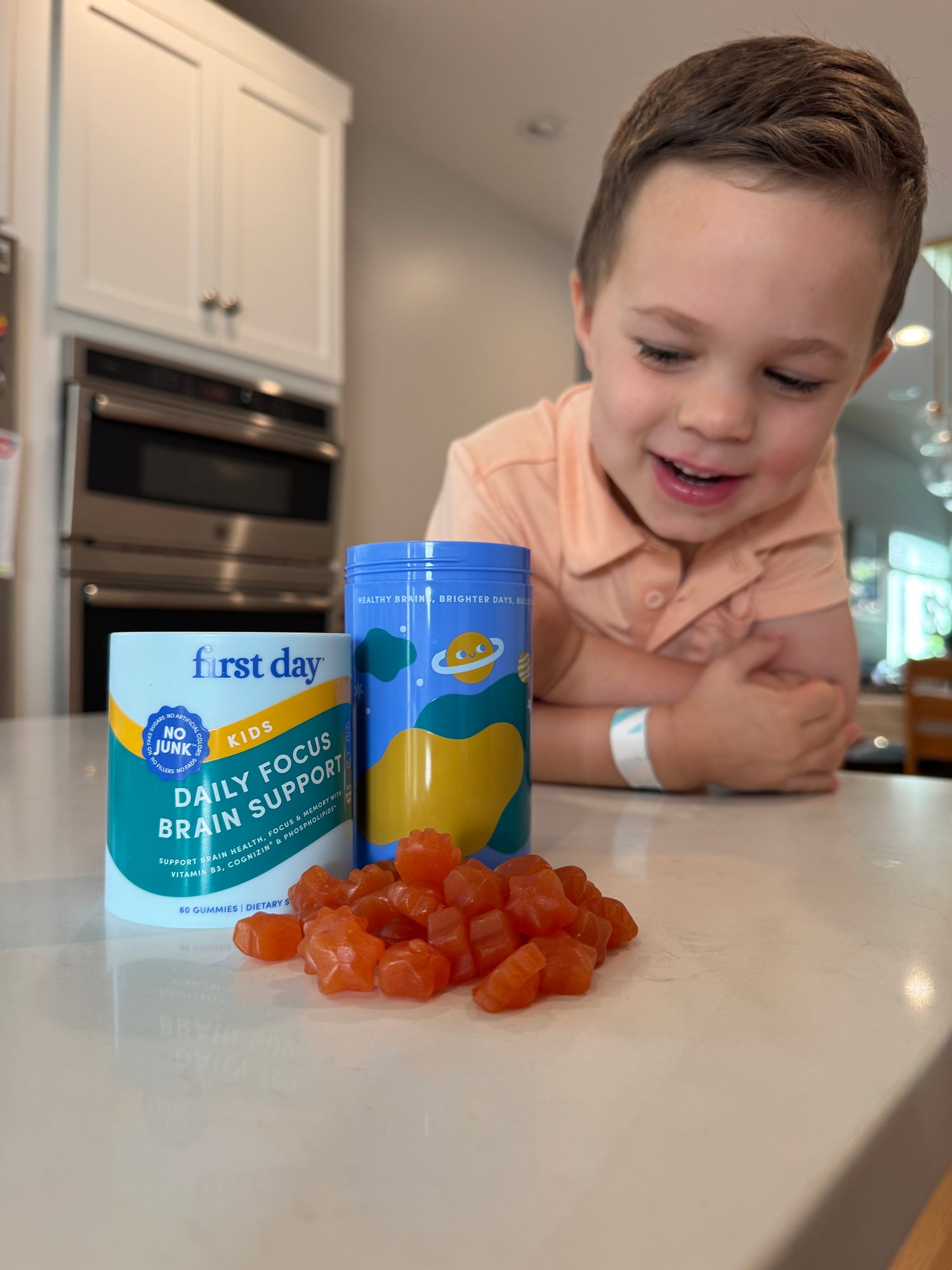A young boy smiles while looking at orange gummy vitamins on a counter next to two containers labeled first day Kids Daily Focus Brain Support. The scene is set in a bright kitchen.