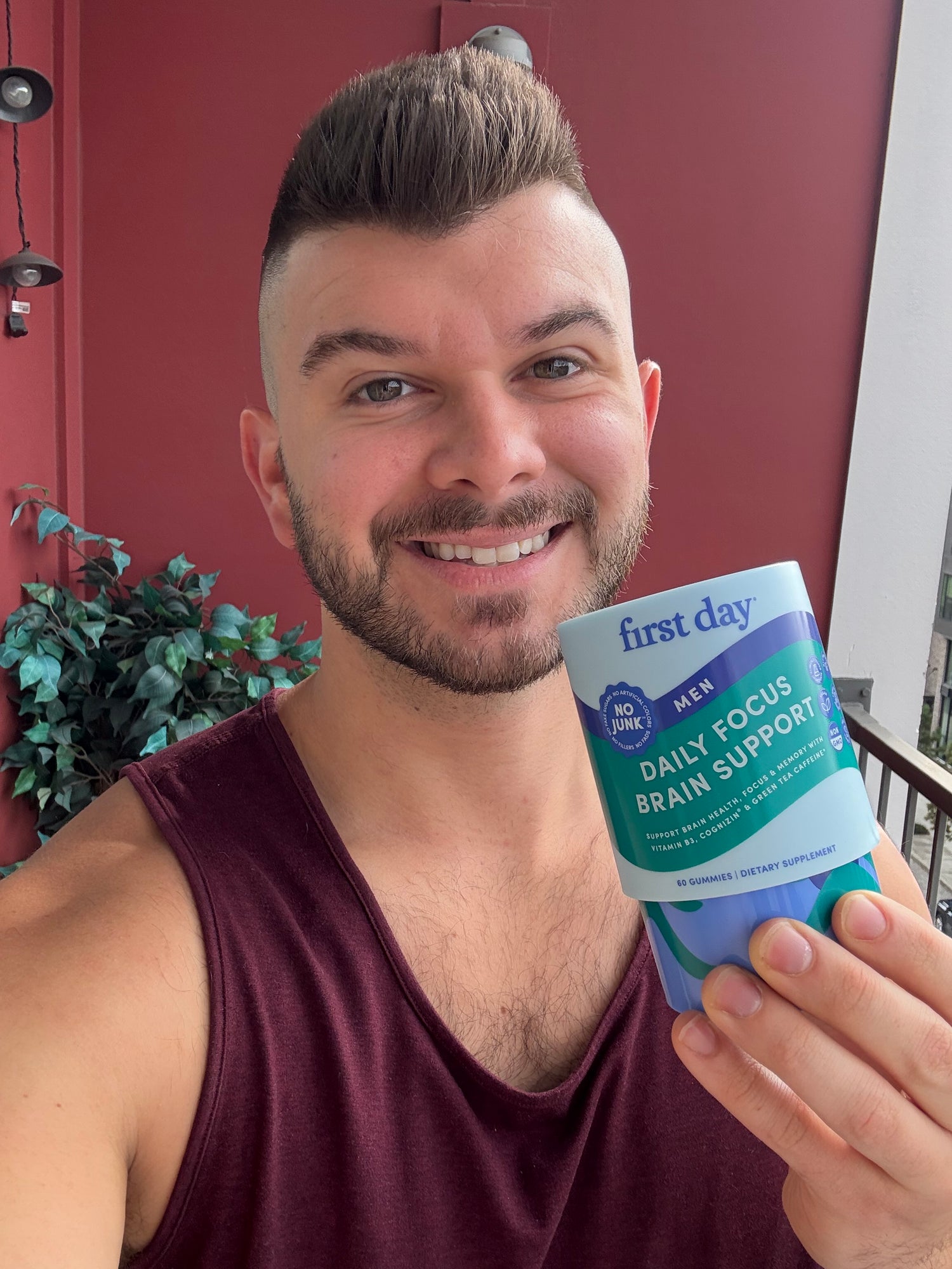 A smiling man with short hair and a trimmed beard holds up a container labeled Daily Focus & Brain Support by First Day, standing on a balcony with a red wall and green plant in the background.