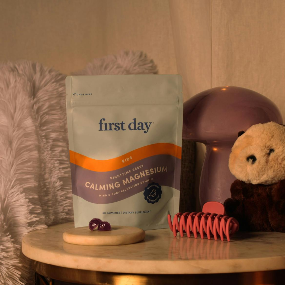 A bag of First Day Calming Magnesium gummies sits on a marble table next to a pink hair clip, a plush toy, a fuzzy pillow, a purple mushroom lamp, and a plate with two gummies.