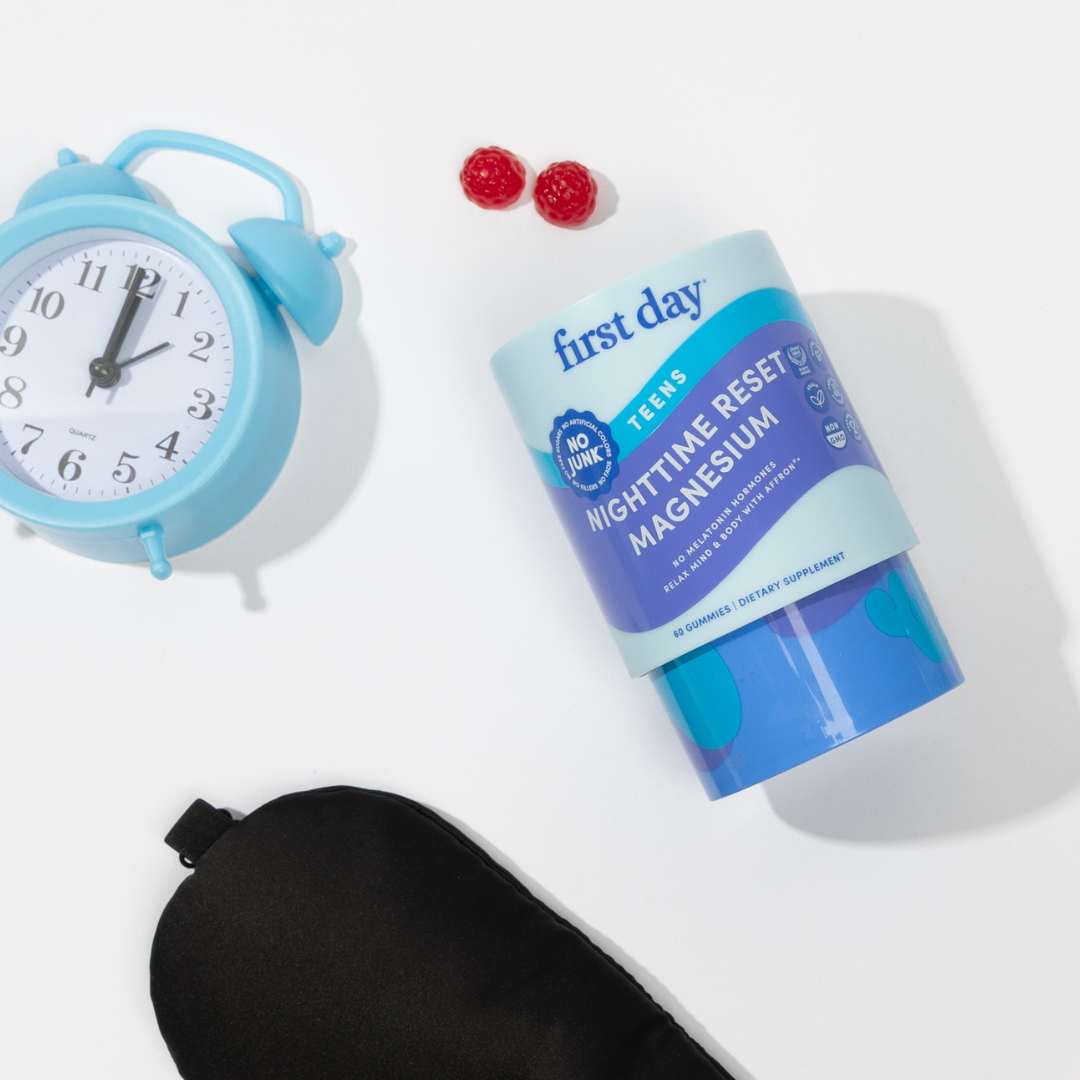 A blue alarm clock, two red gummies, a blue First Day Nighttime Reset Magnesium supplement container, and a black sleep mask are arranged on a white background.