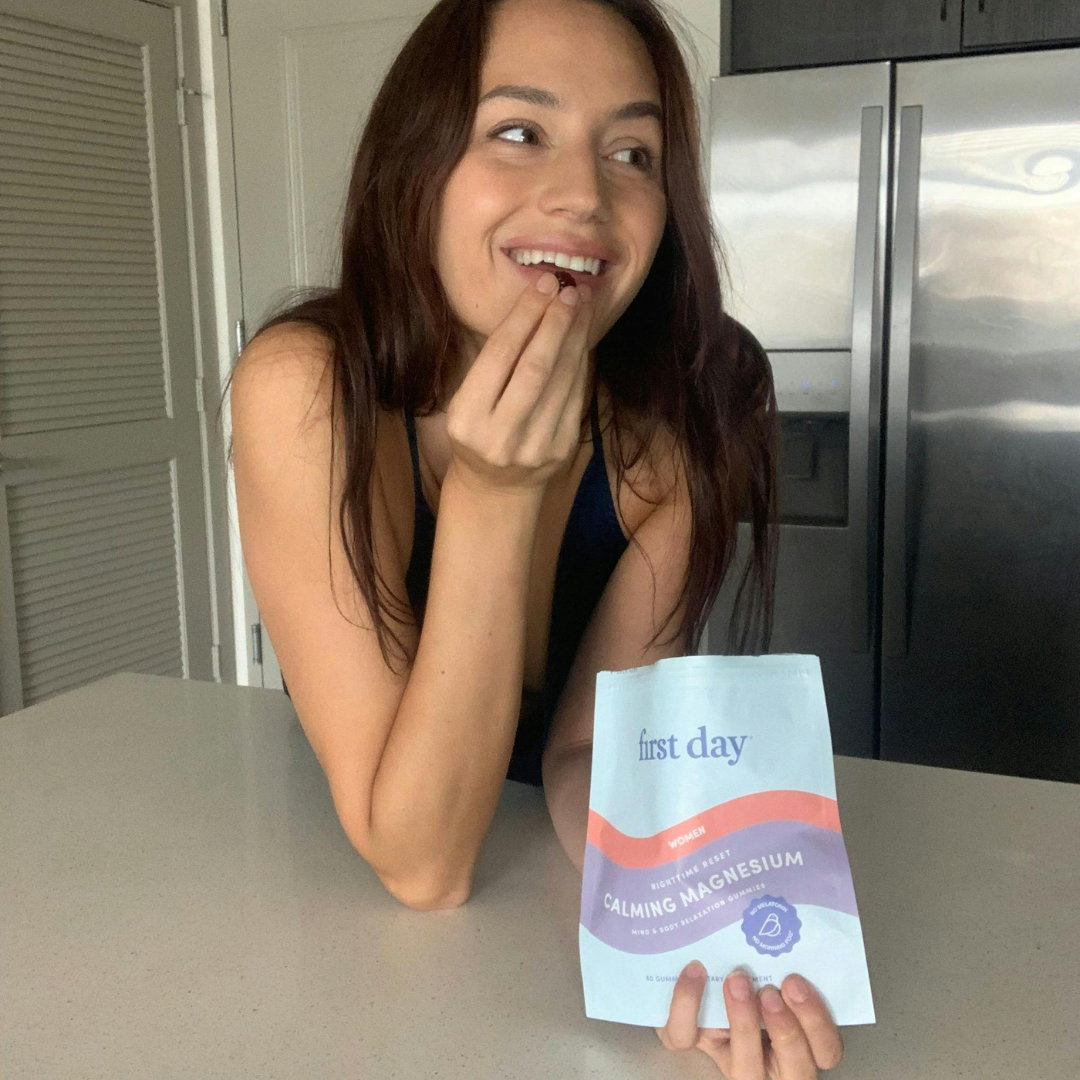 A woman with long brown hair smiles while leaning on a kitchen counter, holding a pouch labeled first day Calming Magnesium and eating a piece of the supplement.