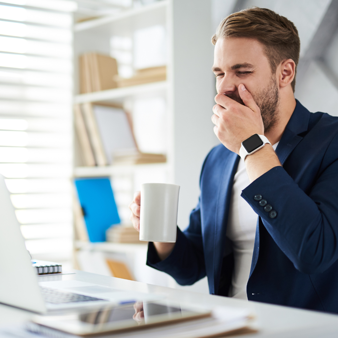 A man in a suit sits at a desk with a laptop, holding a mug and covering his mouth as he yawns. There are shelves with books and binders in the background.