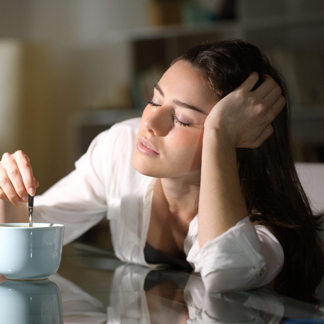 A tired woman sits at a table with her head resting on one hand and her eyes closed, stirring a cup with the other hand, appearing exhausted or sleepy.