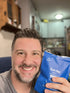A smiling man with short dark hair and a beard holds a blue packet labeled first day MEN’S Daily Provisions MULTI in a kitchen setting.