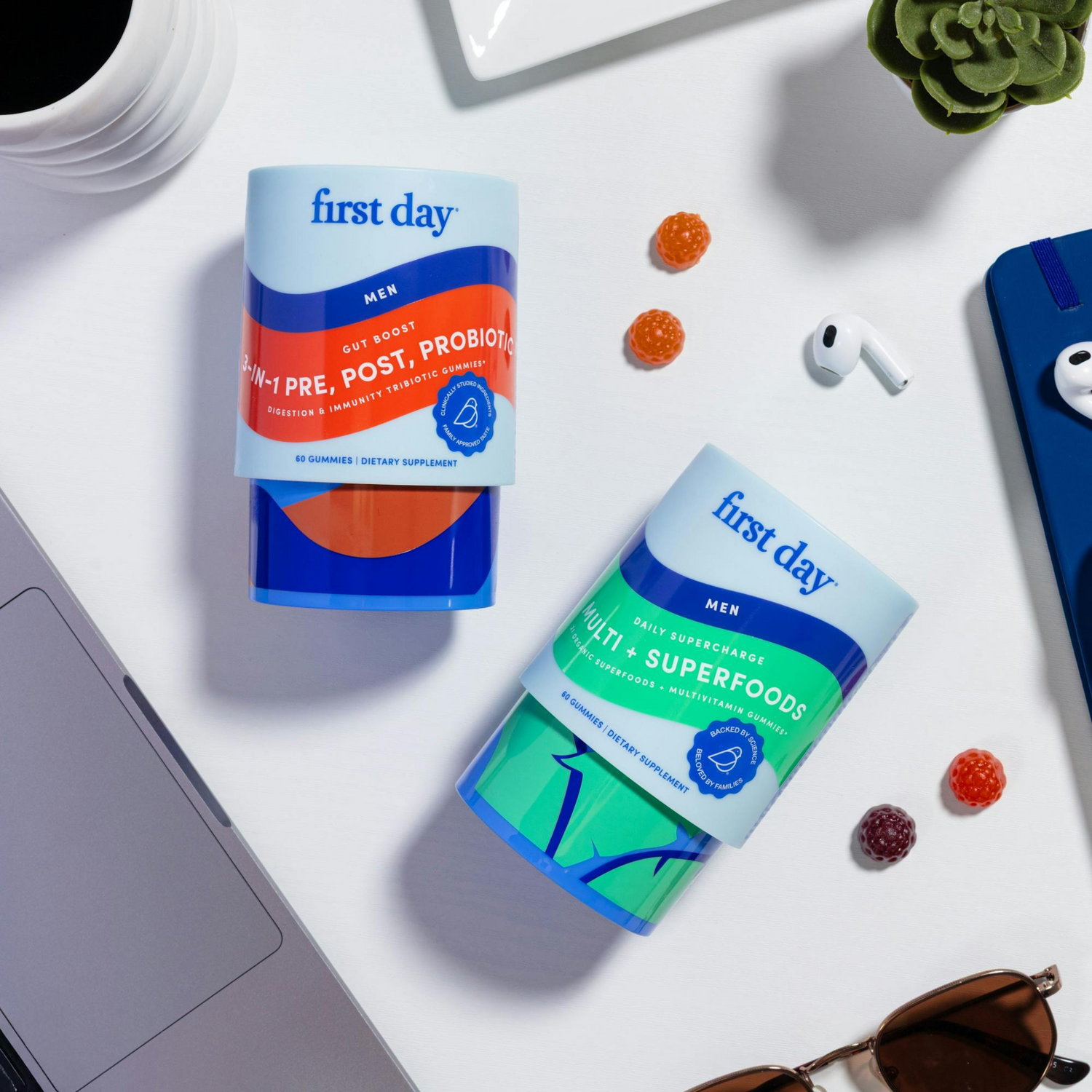 Two containers of First Day mens supplements are on a white surface with a laptop, sunglasses, AirPods, phone, coffee cup, and scattered gummy supplements nearby.