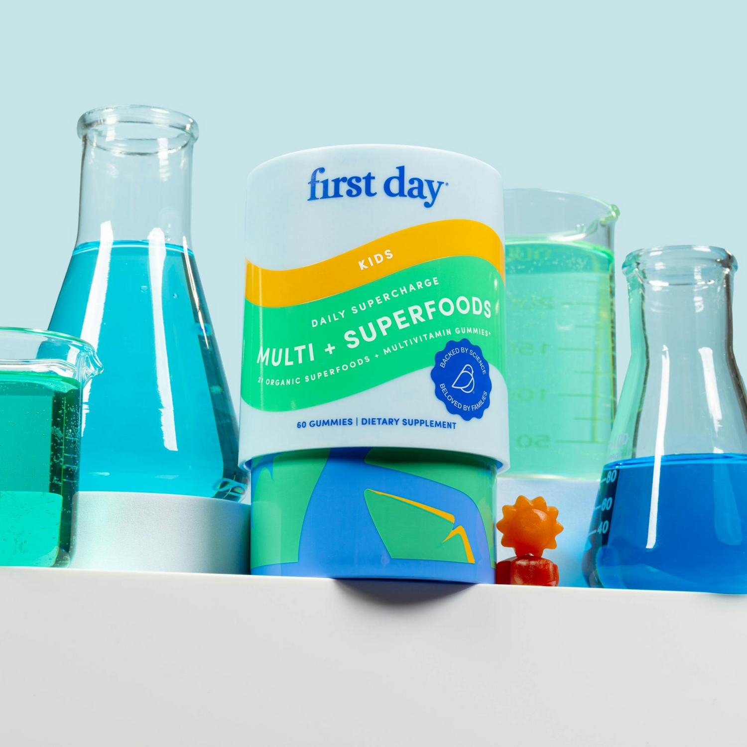 A bottle of First Day Kids Multi + Superfoods gummies sits on a white surface, surrounded by lab beakers filled with blue liquid, against a light blue background. An orange toy and gummy are beside the bottle.