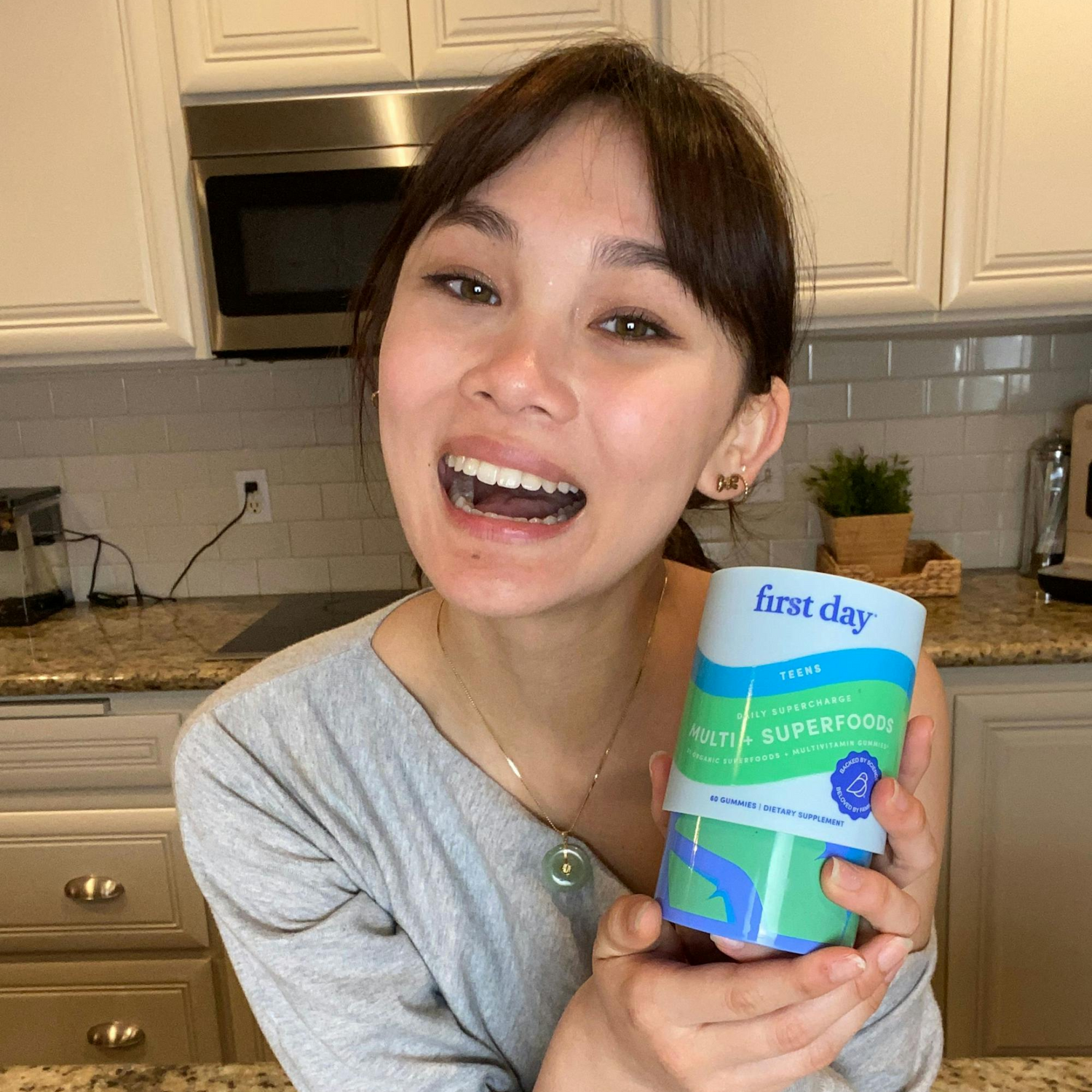 A young woman smiles in a kitchen while holding a container labeled first day Teens Multi + Superfoods. She has dark hair and is wearing a gray shirt. White cabinets and a countertop are visible in the background.