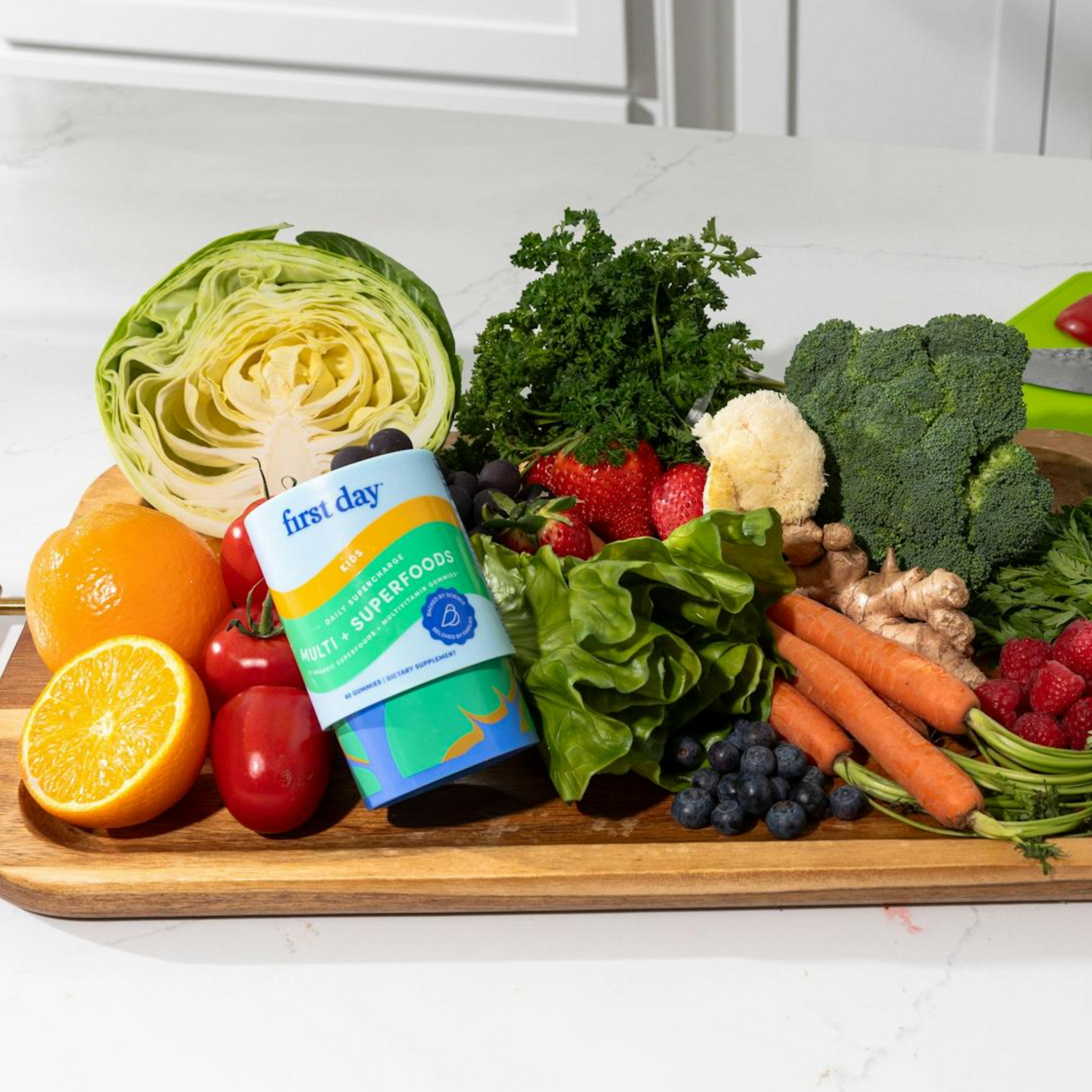A wooden board topped with a variety of fresh fruits and vegetables, including cabbage, carrots, broccoli, lettuce, strawberries, blueberries, tomatoes, an orange half, and a container labeled first day Multis + Superfoods.