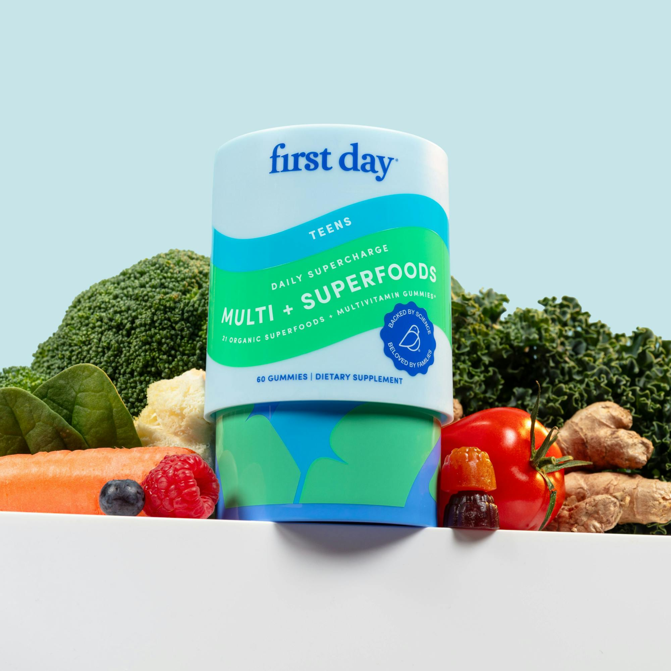 A container of First Day Teens Multi + Superfoods gummies is displayed in front of assorted fresh vegetables and fruits, including kale, tomato, carrot, blueberry, and raspberry, against a light blue background.