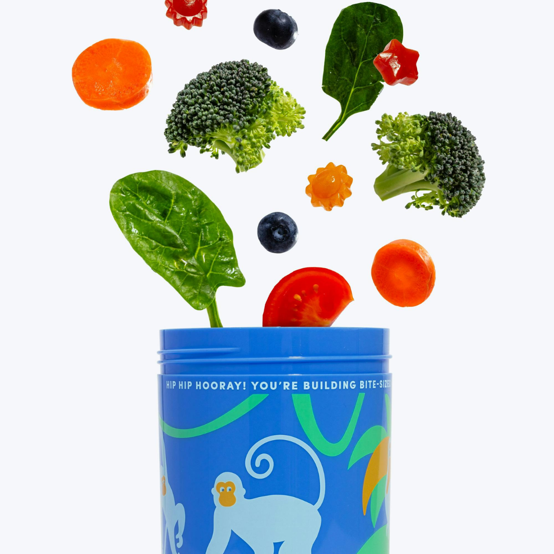 A blue container with a monkey design is open, and assorted vegetables and fruits—including broccoli, spinach, carrot, tomato, blueberry, and bell pepper—are floating above it against a white background.